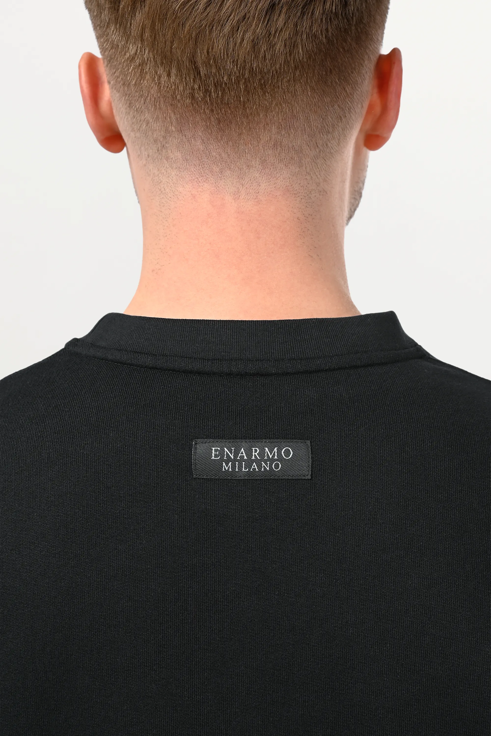 Close-up of tonal black ENARMO woven label at upper back of oversized cotton T-shirt, premium craftsmanship detail on deep black fabric