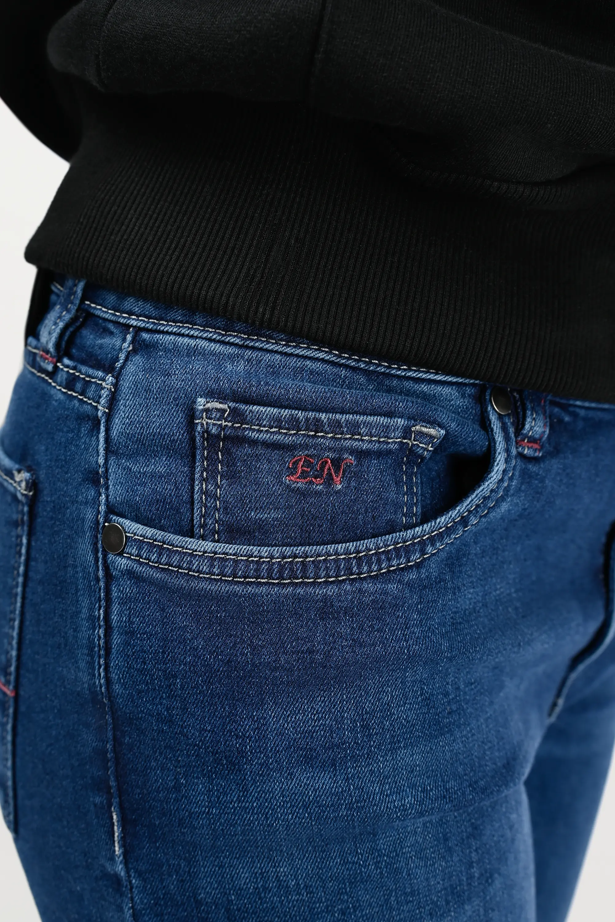 Close-up of coin pocket on mid blue skinny jeans, featuring red bordeaux EN monogram embroidery, signature detail in women’s premium denim.