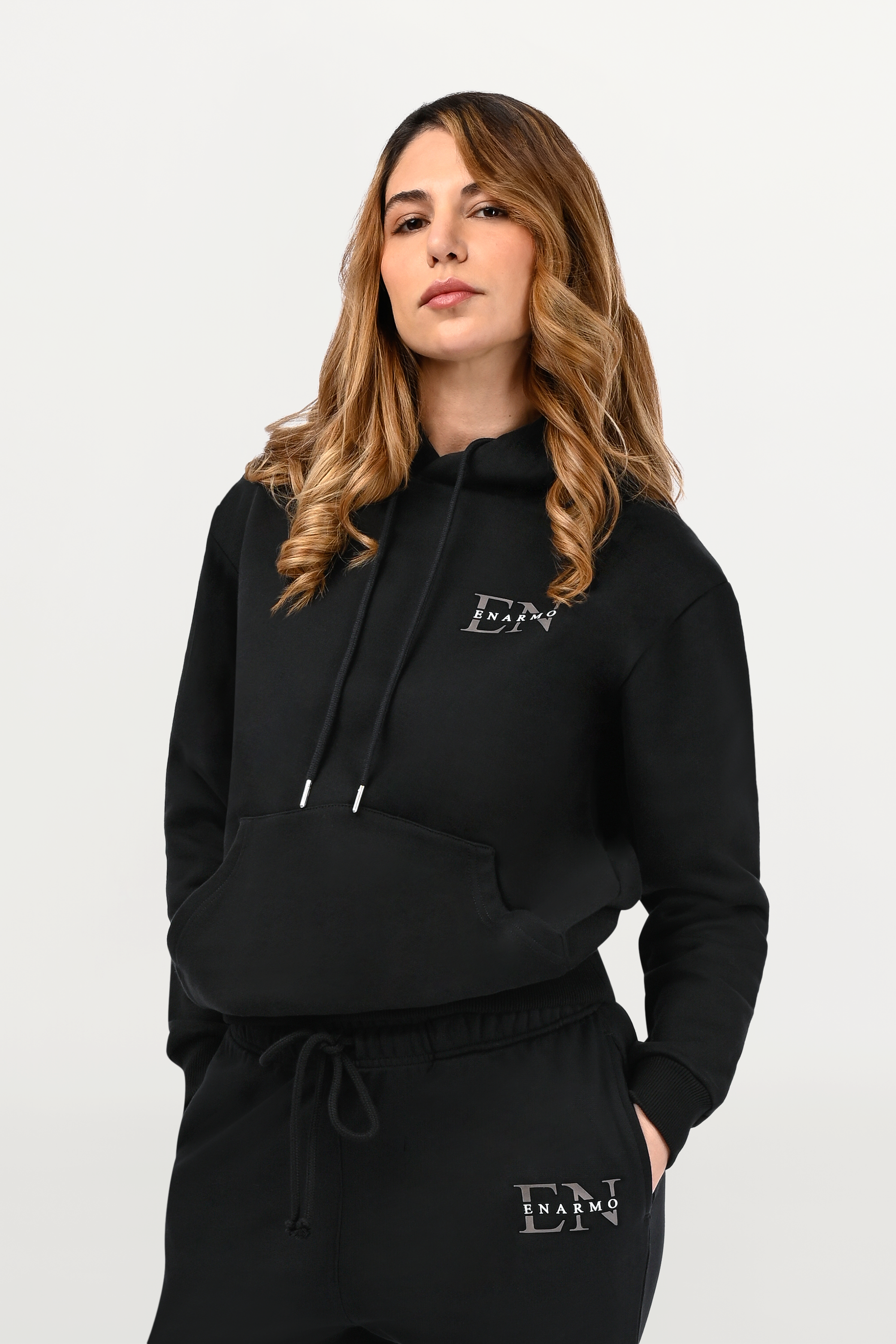 Close-up of chest logo detail on black cotton hoodie, anthracite monogram with subtle white signature accent on premium material.