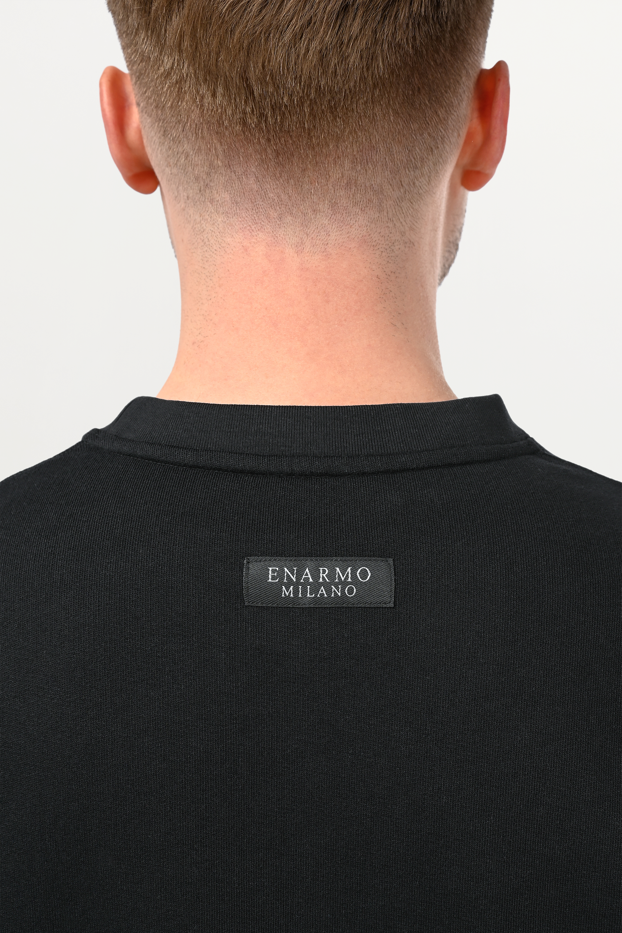 Close-up of tonal black ENARMO woven label at upper back of oversized cotton T-shirt, premium craftsmanship detail on deep black fabric