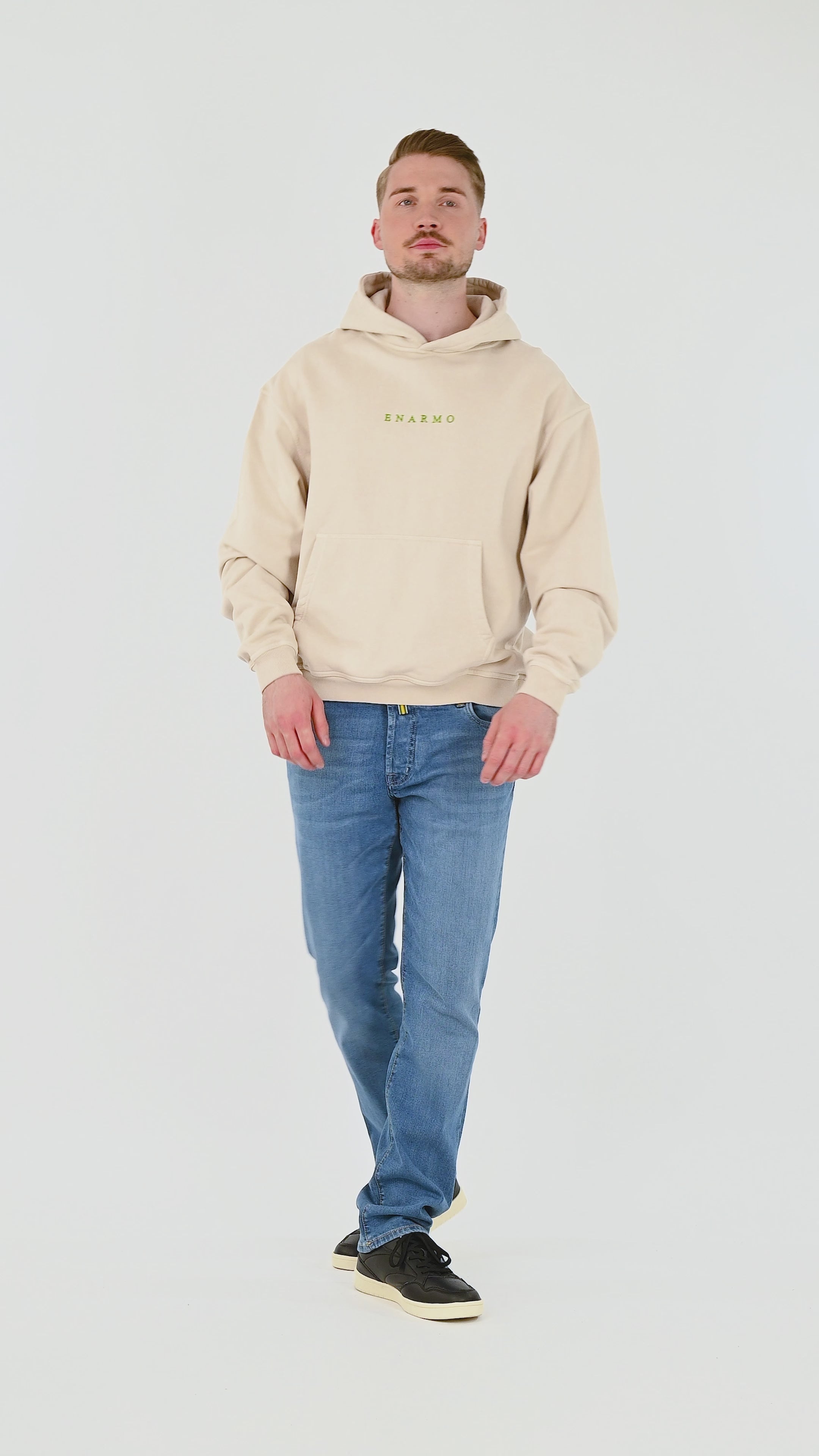 Male model wearing beige oversized cotton hoodie with green embroidery and large back print, turning, posing and moving from front, side and back to highlight fit and silhouette.
