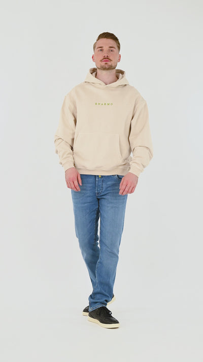 Male model wearing beige oversized cotton hoodie with green embroidery and large back print, turning, posing and moving from front, side and back to highlight fit and silhouette.