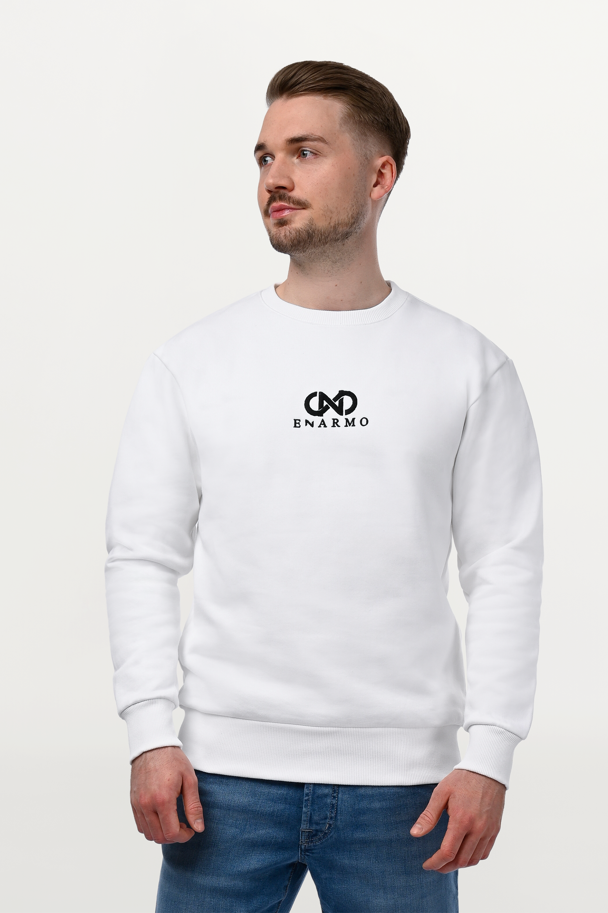 Male model wearing white cotton ENARMO sweatshirt, mid-length view to knee, looking sideways, black embroidered logo detail and clean regular fit silhouette.